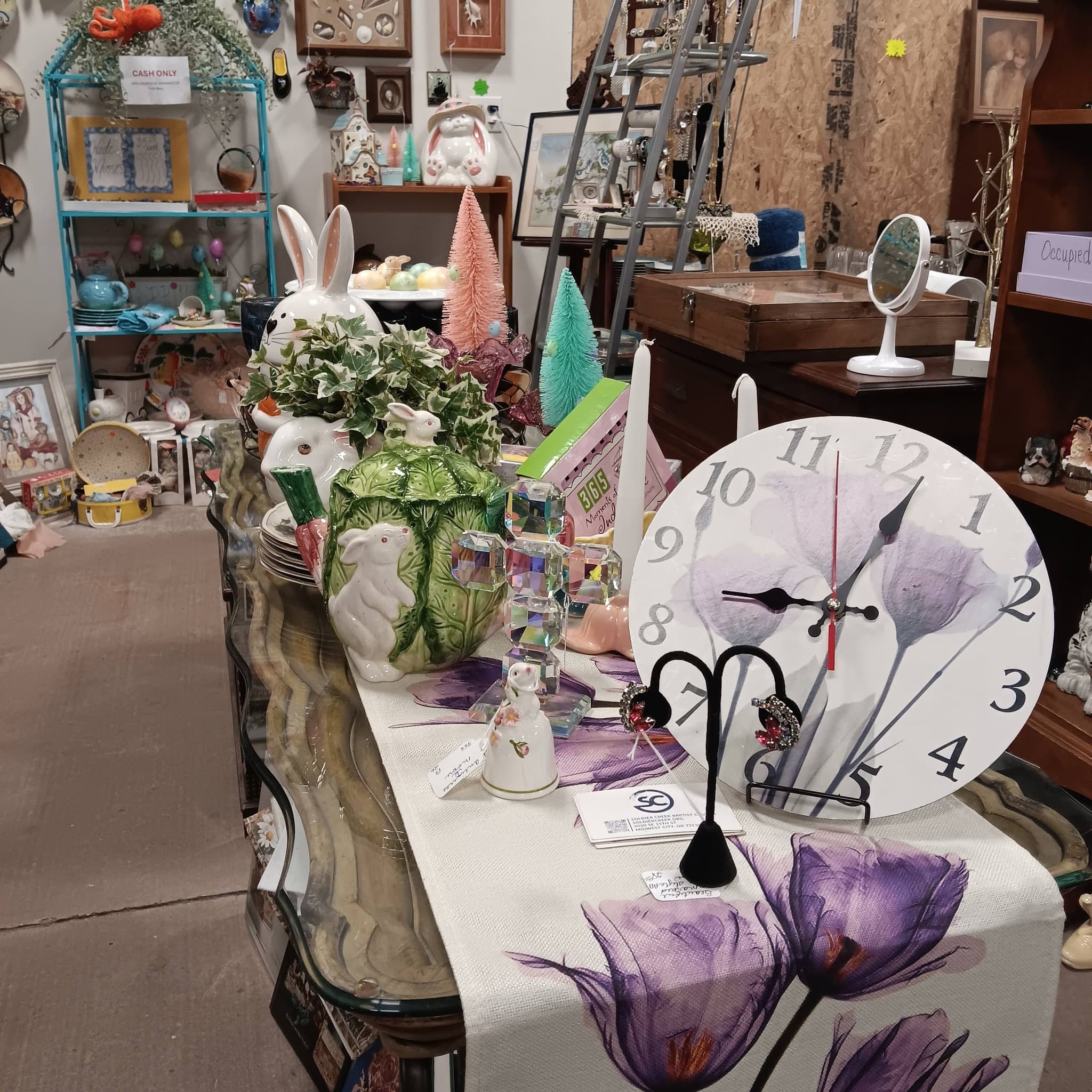 Vendor booth with vintage clock, ceramic rabbit figurine, purple floral tablecloth, and vanity mirror