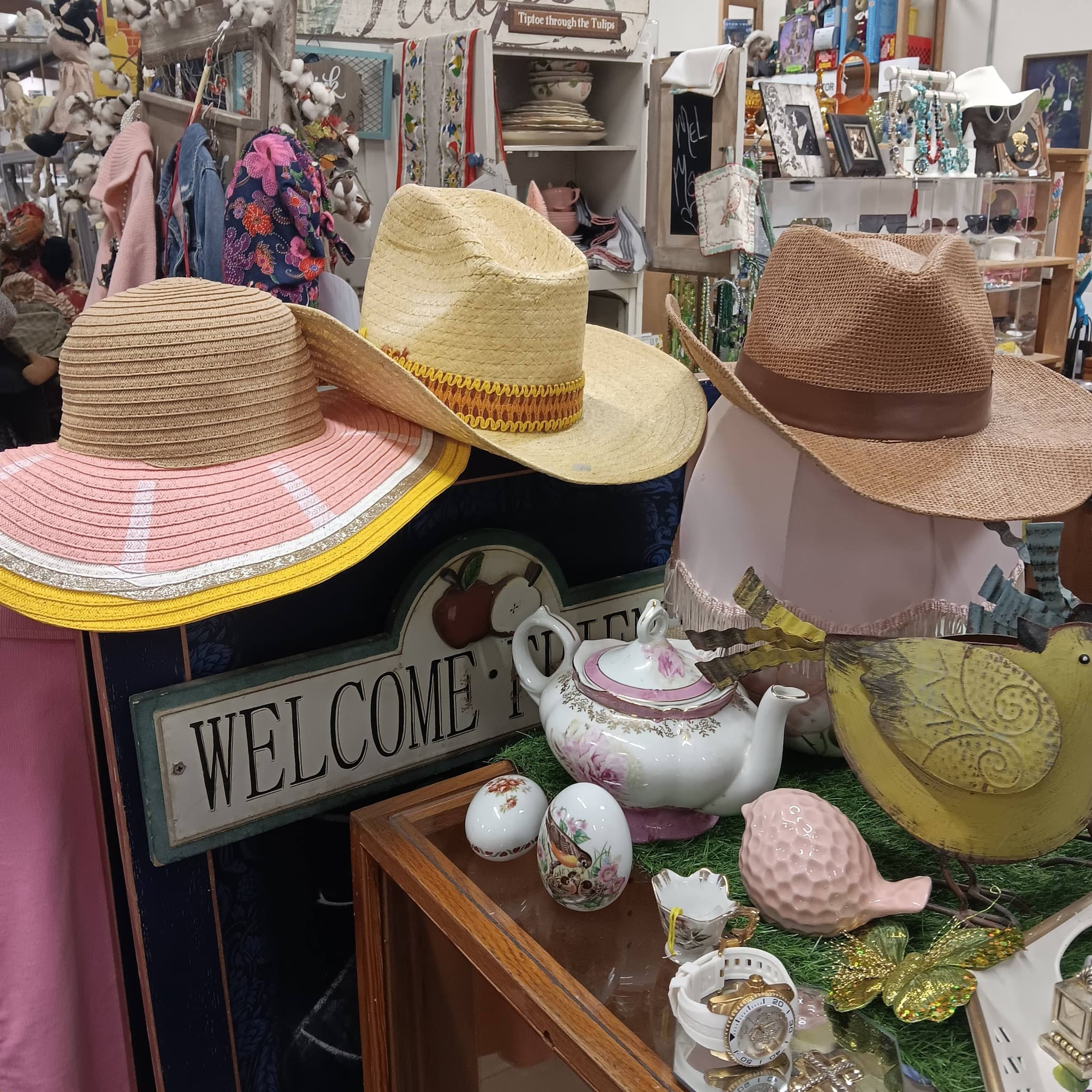 Straw sun hats and cowboy hats above a Welcome sign, teapot, and garden decor